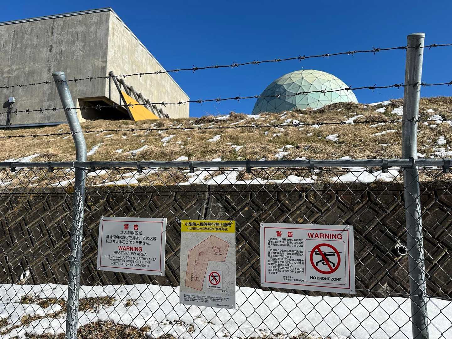 登山道が自衛隊のレーダー基地に突き当たります。有刺鉄線が張り巡らされ、入っちゃダメ、ドローンを飛ばすな、と書いてあります。
後方はレーダードームです。中でレーダーが回っており、その回転音が唸りのように聞こえています。